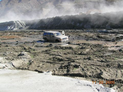 1985 Ford Bronco - A tan SUV is stuck in deep mud, its driver's side door ajar. The scene appears to be a construction or demolition site, with heavy machiner…