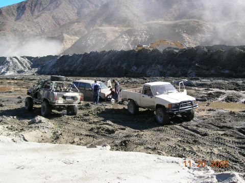 1985 Ford Bronco - Three vehicles are stuck in a muddy, off-road area with construction equipment in the background.
