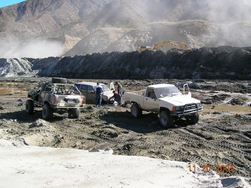 Tan pickup truck with lifted suspension and open door, parked in mud next to a tan SUV and a camouflaged off-road vehicle