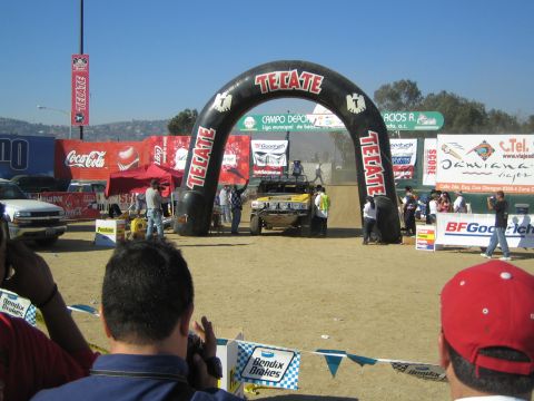 1990 Ford Bronco - spectator crowd watches an off-road race truck pass beneath an inflatable Tecate arch on a dirt course lined with Coca-Cola and BFGoodrich…