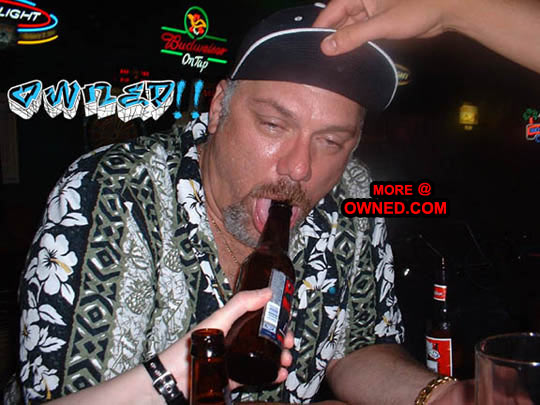 Man with a black baseball cap and floral shirt drinks from a beer bottle in a bar with neon signs