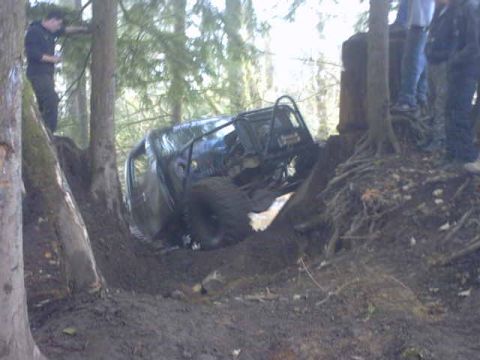 1995 Ford Bronco - An off-road vehicle is stuck in a muddy ditch surrounded by trees, with onlookers standing on a bank above.