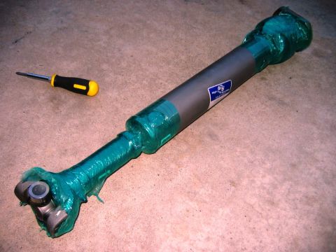1985 Ford Bronco - A driveshaft, wrapped in protective green plastic, rests on a concrete floor next to a screwdriver.