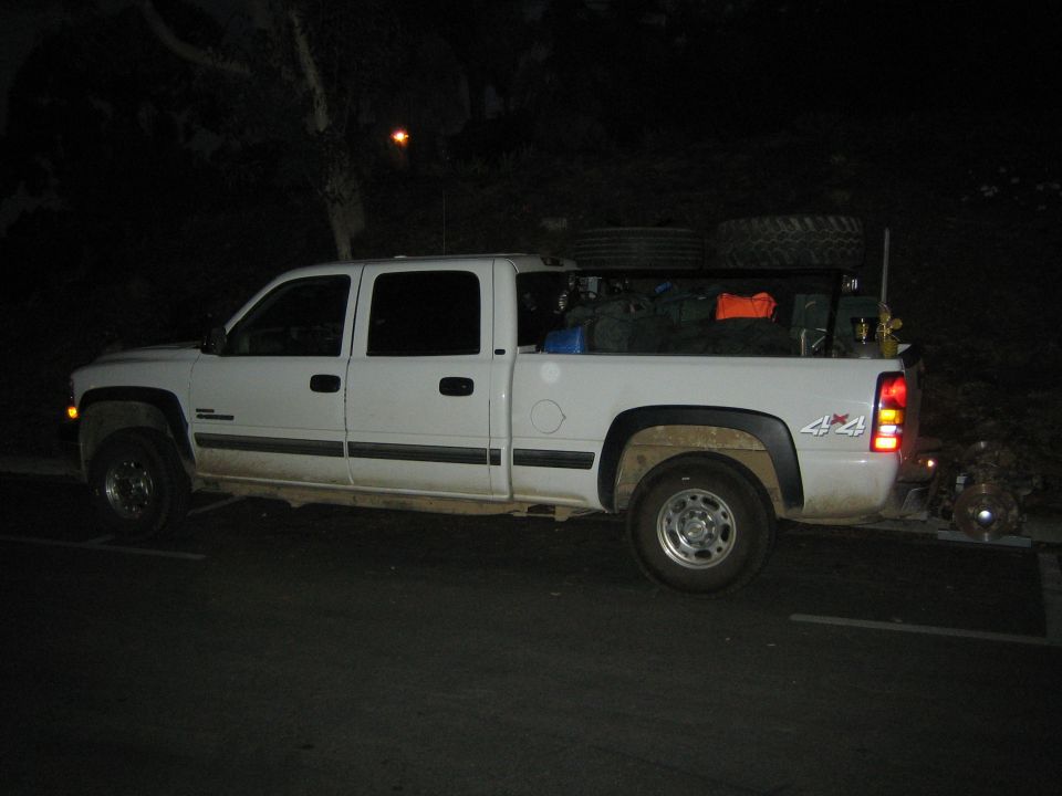White crew-cab 4x4 pickup parked at night with bed loaded with gear and spare tires