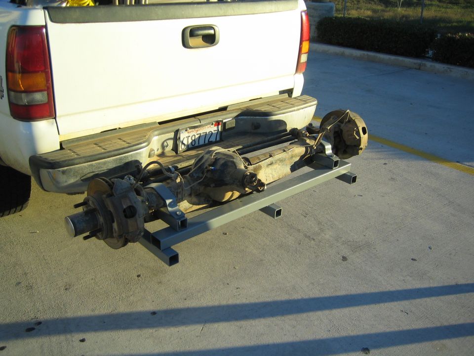 Vehicle axle and driveshaft assembly on a metal rack behind a white pickup tailgate
