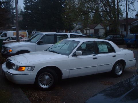 1999 Ford Crown Victoria - A white sedan is parked on a street with other vehicles and houses visible.