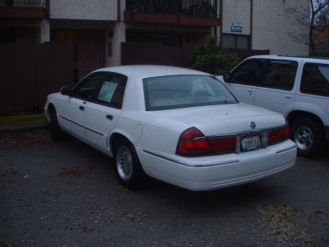 1999 Ford Crown Victoria - A white sedan is parked in a lot next to a white SUV, with a building in the background.
