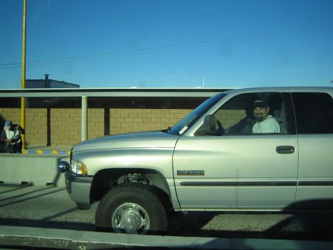1990 Ford Bronco - silver Dodge Ram pickup with a bearded, smiling driver travels alongside a low brick building under blue sky.