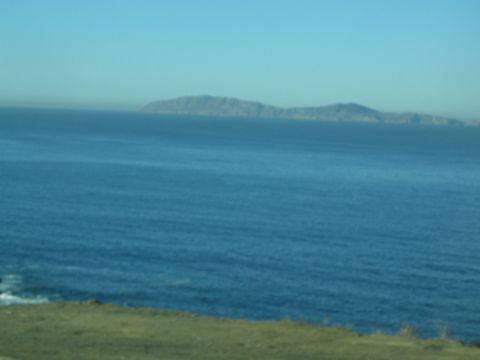 1990 Ford Bronco - hazy island rises across a calm blue ocean, viewed from a grassy coastal bluff under clear sky.