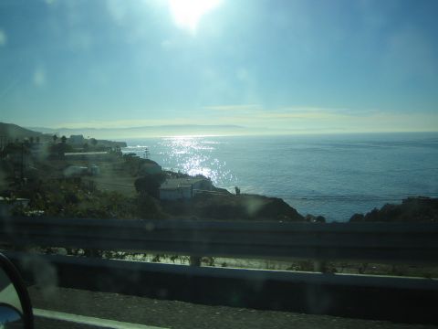 1990 Ford Bronco - Sun glints off the ocean beyond a coastal hillside dotted with buildings, framed by a highway guardrail and car mirror.