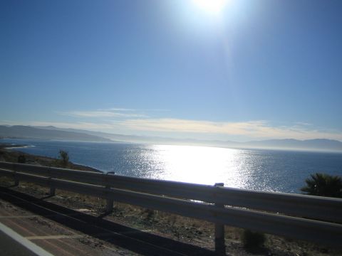 1990 Ford Bronco - sun blazes high over a calm ocean bay and distant coastal hills, framed by a roadside guardrail.