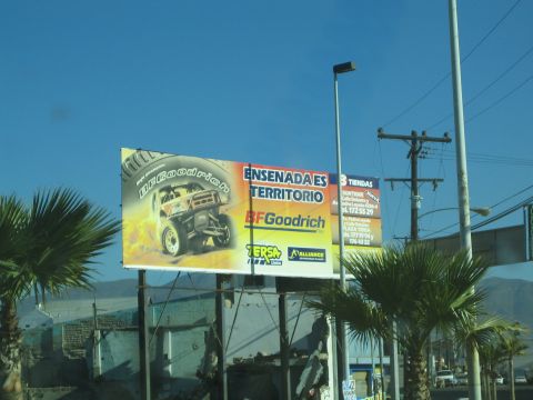 1990 Ford Bronco - yellow roadside billboard reads Ensenada es Territorio BFGoodrich beside palm trees and power lines under blue sky.
