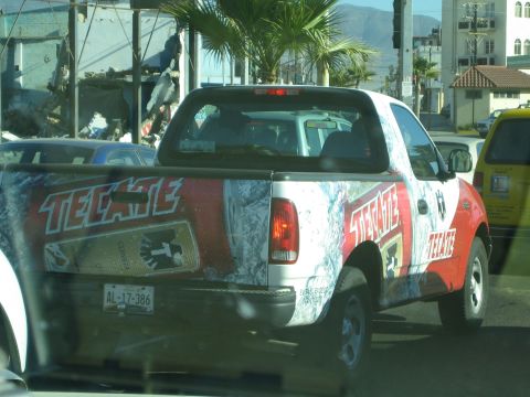 1990 Ford Bronco - pickup wrapped in Tecate beer livery sits in traffic beside palm trees and buildings with mountains beyond.