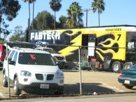 1990 Ford Bronco - black and yellow Fabtech race trailer with flame graphics and Toyo Tires logo sits behind a chain-link fence beside a white Pontiac minivan.