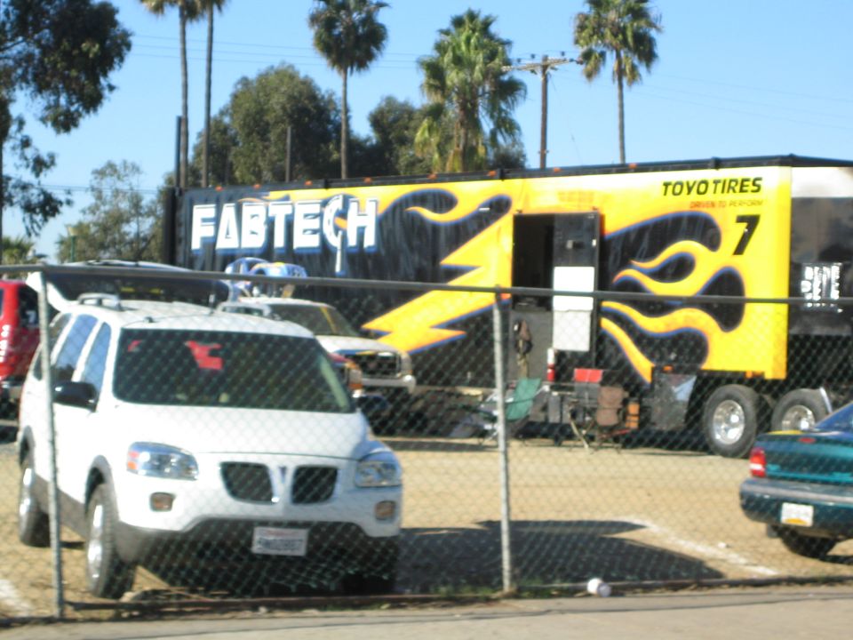 Black and yellow Fabtech race trailer behind a chain-link fence with a white minivan