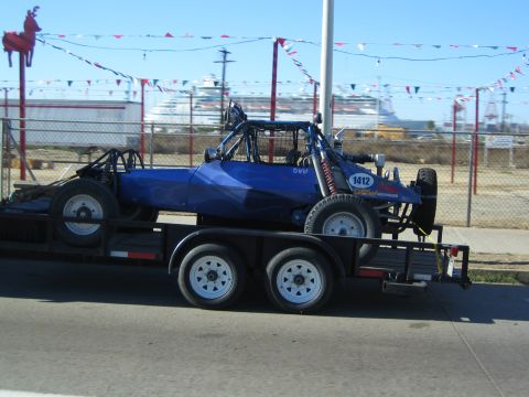 1990 Ford Bronco - blue tube-frame off-road race buggy marked 1412 rides on a flatbed tandem-axle trailer, with a cruise ship docked behind a fence.