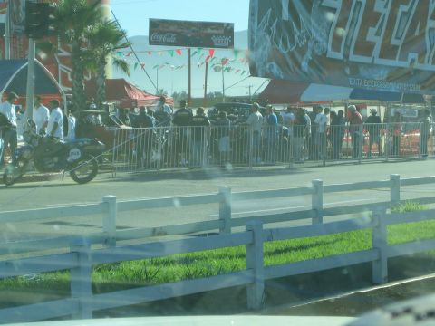 1990 Ford Bronco - crowd gathers behind metal barriers at a street event with a motorcycle, vendor tents and a Coca-Cola sign, seen through a car window.