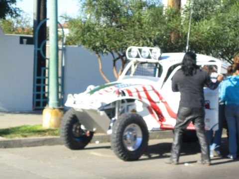 1990 Ford Bronco - white tube-frame off-road buggy painted with a U.S. flag motif is parked on a street as people stand beside it.