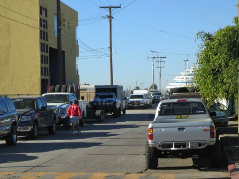 1990 Ford Bronco - Trucks and trailers line both sides of a street leading toward a large docked cruise ship, with a person in a red jacket walking by.