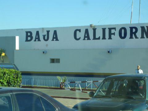 1990 Ford Bronco - white building bears large painted Baja Californ lettering above a fenced lot where dark cars are parked and a man stands.