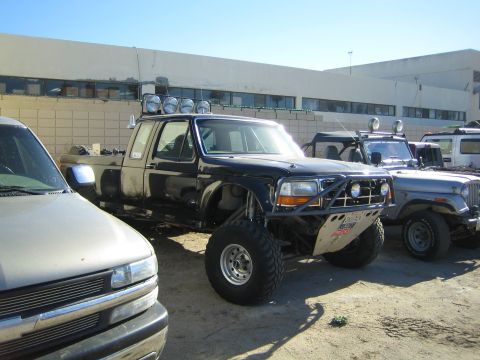 1990 Ford Bronco - lifted black 1990s Ford crew-cab pickup on oversized off-road tires with a roof-mounted light bar sits parked beside other vehicles.