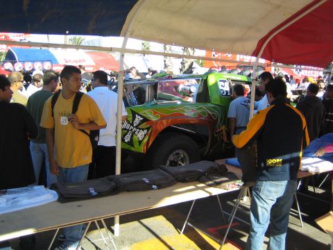 1990 Ford Bronco - green off-road race truck with orange flames is displayed under a tent while a crowd browses merchandise at an outdoor event.