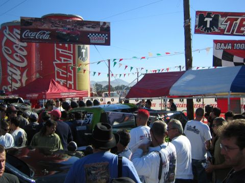 1990 Ford Bronco - dense crowd gathers among race vehicles at a Baja 1000 contingency event beneath Coca-Cola and Tecate banners.