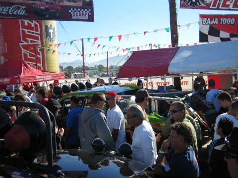 1990 Ford Bronco - Spectators pack in around off-road race vehicles beneath Tecate and Baja 1000 banners at a sunny outdoor event.