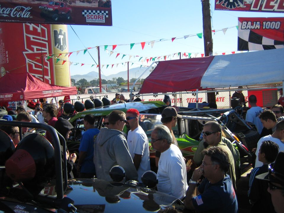 Spectators crowd around race vehicles under Tecate and Baja 1000 banners at an outdoor desert race event
