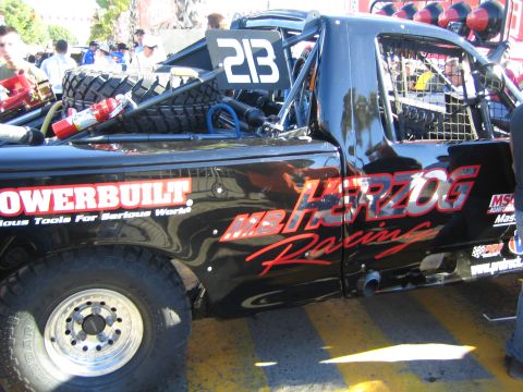 1990 Ford Bronco - black race truck marked number 213 with Powerbuilt and Herzog Racing graphics carries spare tires in its bed at an event.