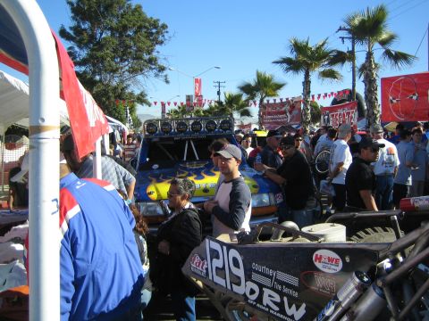 1990 Ford Bronco - blue-and-yellow race truck and a number 1219 buggy are displayed among event crowds beneath palm trees and a clear sky.