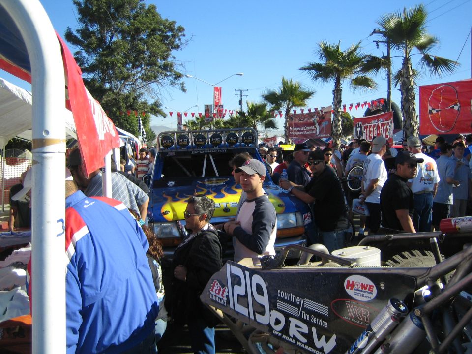 Blue and yellow off-road race truck and a number 1219 buggy displayed among crowds under palm trees and blue sky
