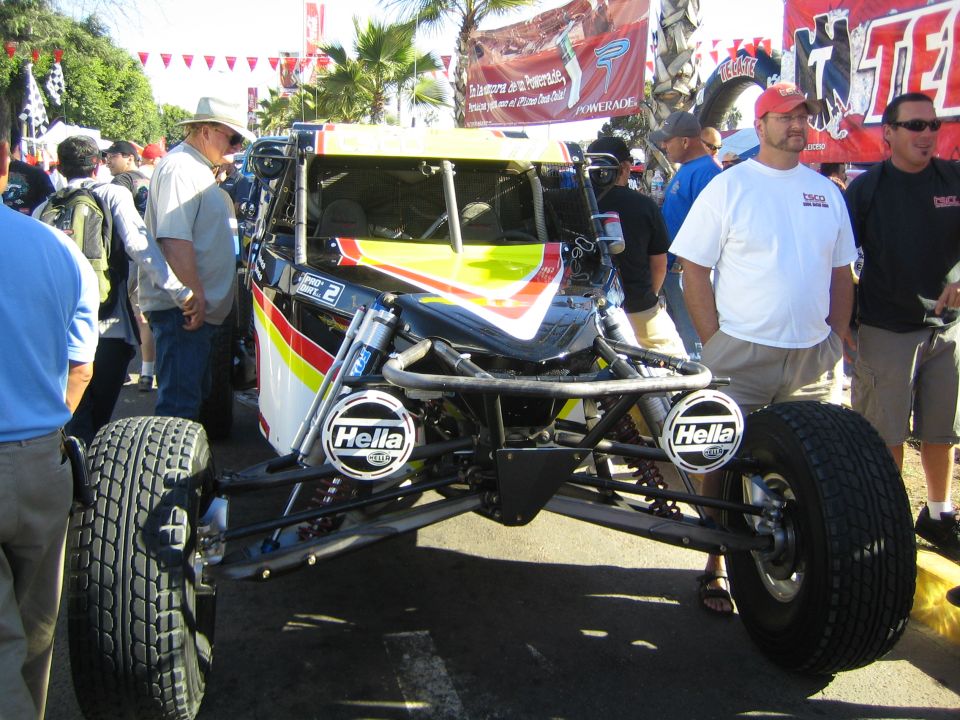 Open-wheel off-road race car chassis with Hella lights and exposed suspension on display in a crowd