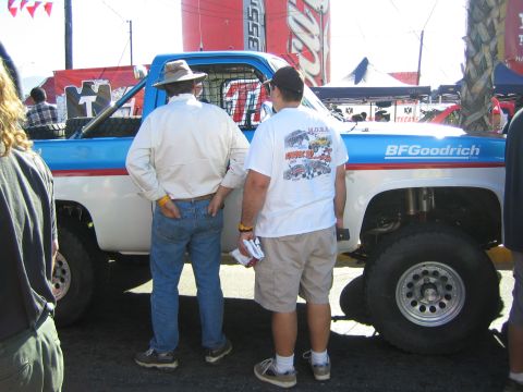 1990 Ford Bronco - Two men lean in to inspect a blue-and-white lifted off-road race truck bearing BFGoodrich graphics at an outdoor event.
