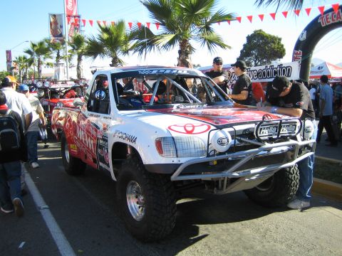 1990 Ford Bronco - red-and-white Toyota trophy truck with Yokohama and Hella branding is staged under palm trees as crew work around it at an event.