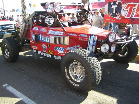 1990 Ford Bronco - red open-wheel off-road race buggy decked with Fox, BFGoodrich and Truss sponsor graphics sits on pavement at a race event.