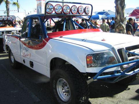 1990 Ford Bronco - white-and-red off-road race truck lettered Mango Racing with a six-lamp roof light bar sits on display at an outdoor event.