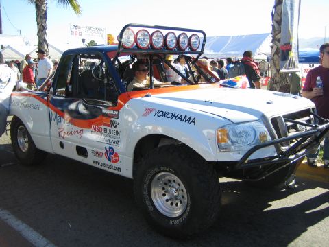1990 Ford Bronco - driver sits inside a white off-road race truck with orange graphics, Yokohama branding and a roof light bar at a race event.