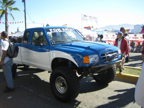1990 Ford Bronco - blue-and-white lifted Ford extended-cab pickup on oversized tires displays a FOR SALE windshield banner at an outdoor event.