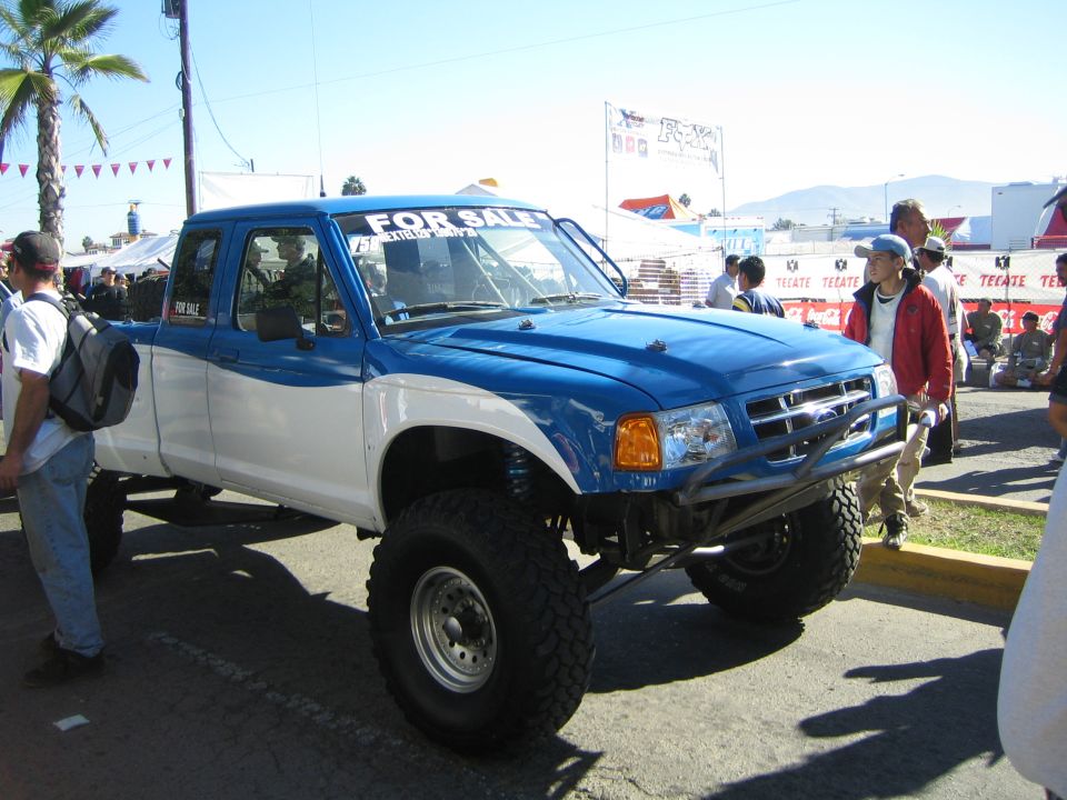 Blue and white lifted Ford extended-cab pickup with a FOR SALE windshield banner on big tires at an event