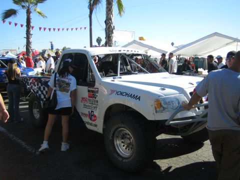 1990 Ford Bronco - Spectators gather around a white off-road race truck carrying Yokohama and MSD graphics beneath palm trees and a clear sky.
