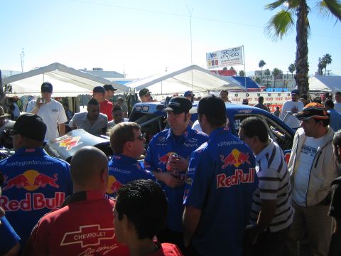 1990 Ford Bronco - crew wearing blue Red Bull and Chevrolet racing shirts gathers to talk near tents at an outdoor desert race event.