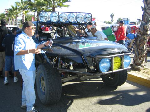 1990 Ford Bronco - man photographs a black off-road race truck fitted with a multi-lamp roof bar and yellow driving lights at an outdoor event.