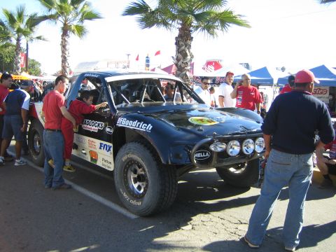 1990 Ford Bronco - red-shirted crew works around a black off-road race truck with BFGoodrich, Hella and Fox graphics beneath palm trees at an event.