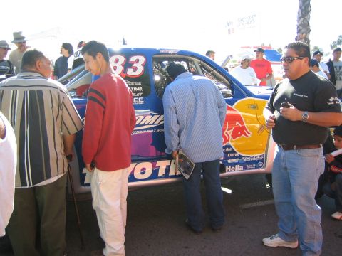 1990 Ford Bronco - Spectators inspect a blue number 83 Toyota off-road race car with USA-themed and Hella graphics at an outdoor race event.