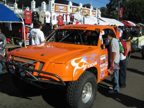 1990 Ford Bronco - orange number 288 off-road race truck with white flame graphics and Yokohama branding sits near merchandise tents at an event.