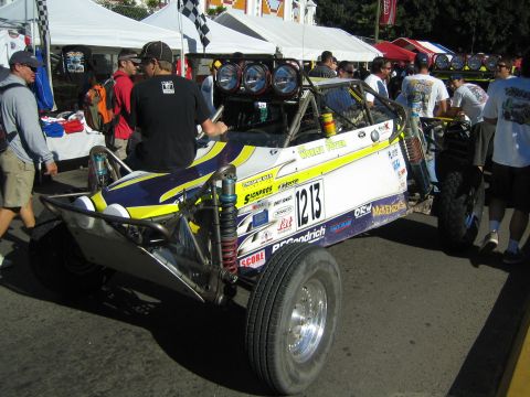 1990 Ford Bronco - blue and yellow tube-frame off-road race buggy, number 1213, parked among crowds and tents at an outdoor racing event.