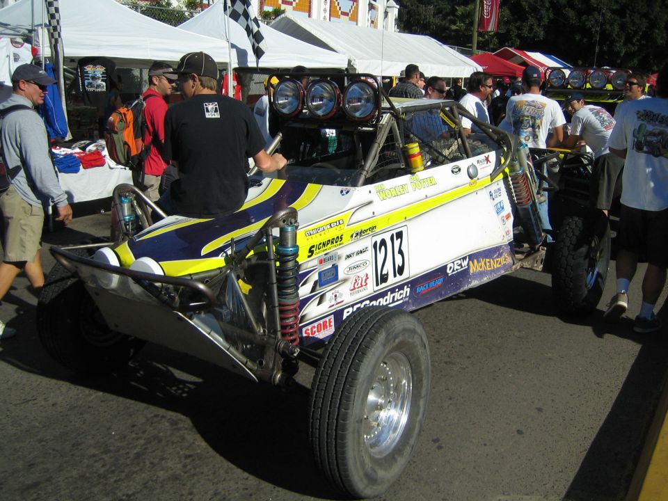 Blue and yellow off-road race buggy number 1213 with overhead light bar at a busy event