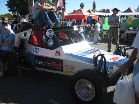 1990 Ford Bronco - white tube-frame off-road buggy, number 1210, with people seated on top, parked in a crowded street lined with tents.