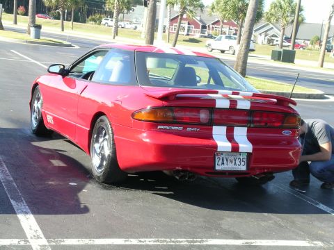 1993 Ford Probe - A red 1993 Ford Probe with white racing stripes is viewed from the rear, with a person checking the bumper.